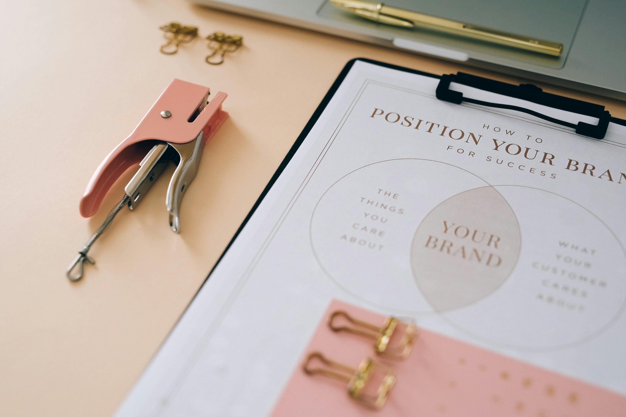A flat lay image of a stylish, feminine workspace featuring a pink stapler, gold binder clips, a clipboard with a business plan worksheet titled "How to Position Your Brand for Success," and a gold pen resting on a silver laptop. The Power of a Well-Written Business Plan.